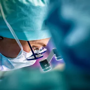 Closeup of a doctor wearing a mask and hat and looking through glasses and magnifying instruments during a surgery.