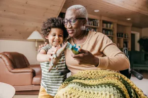 Grandmother in wheelchair playing with dinosaur toys with laughing grandson at home.