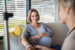 A pregnant patient receives antibiotic treatment through an IV bag in the comfort of home while a friend sits by during treatment.