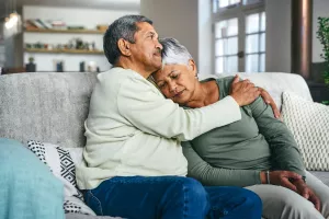 Senior couple sitting on couch together while one partner comforts the other partner who suffers from a mood disorder (depression).