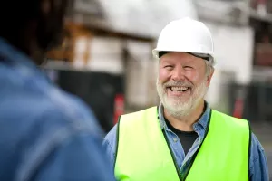 Smiling senior construction worker in green vest and a hard hat talking to coworker on job site.