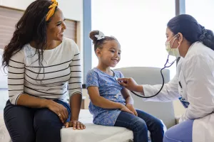 Pediatric patient getting their heart examined with a stethoscope by a doctor while sitting on hospital bed with parent. 